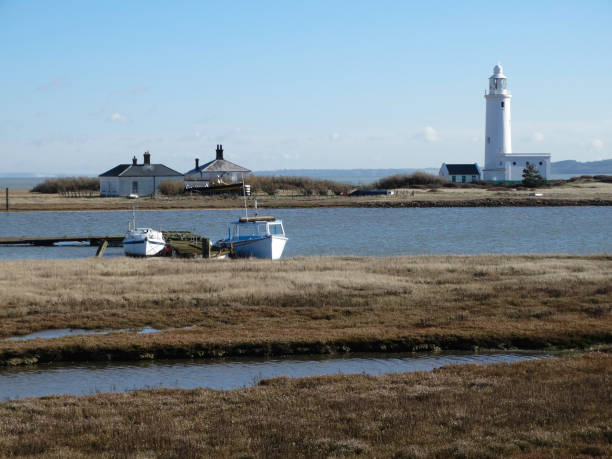 Hurst Point Lighthouse showing both keepers cottages and the out buildings shown across the Lake with two boats moored in the foreground.