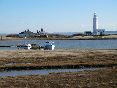 Hurst Point Lighthouse showing both keepers cottages and the out buildings shown across the Lake with two boats moored in the foreground.