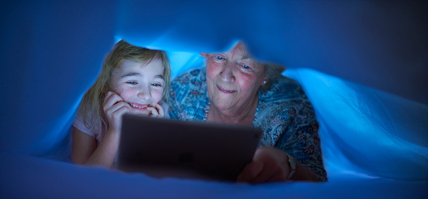 a little girl reads a bedtime story to her gran undr the duvet , using a digital tablet . They are both laughing and having fun .
