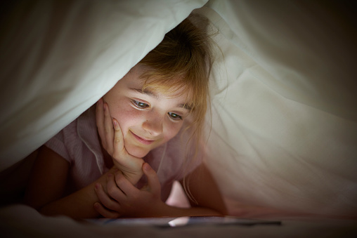 a little girl reads a bedtime story  under the duvet , using a digital tablet .
