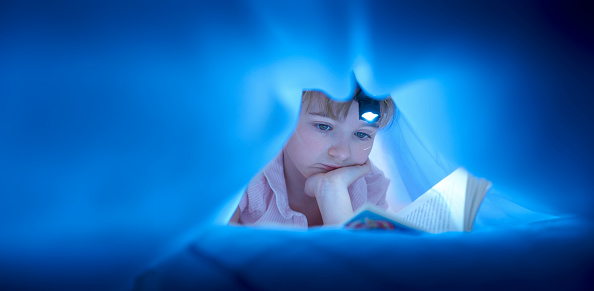 a little girl lies beneath her duvet with a mountaineering head torch on her head shining on her reading book .