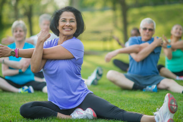 um grupo de adultos idosos estão tomando uma aptidão calss fora no parque. eles estão sentados na grama e estão esticando juntos. - senior couple senior adult action retirement - fotografias e filmes do acervo