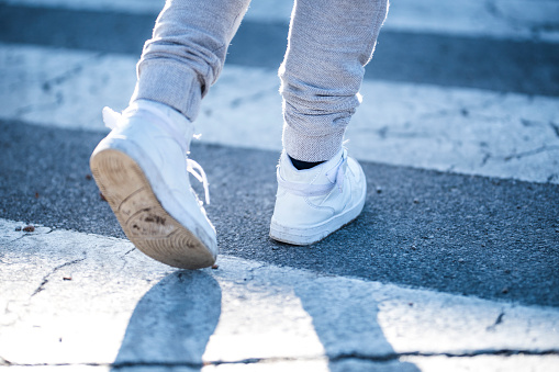 Close up photo of child's legs in white sneakers and grey trousers, crossing the street on a pedestrian crossing in a daytime.