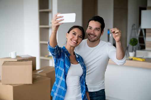 Happy Latin American couple taking a selfie holding keys to their new house and smiling - real estate concepts