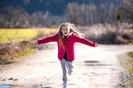 Cute little girl with long blond hair, wearing white sneakers, grey pants and red cardigan, running in nature with wide opened arms and a smile on her face. There is a blue sky and dark wood in the background on a sunny spring day.