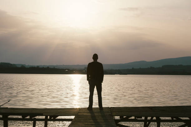 istock Lonely man standing on old wooden lake dock looking at sunset 646714196