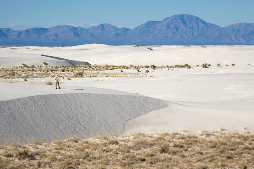 Hiking the rippled, white gypsum sand dunes, a woman hiker with a pony tail walks over the Tularosa Basin in the Chihuahuan Desert with the San Andres Mountains in the distance.
