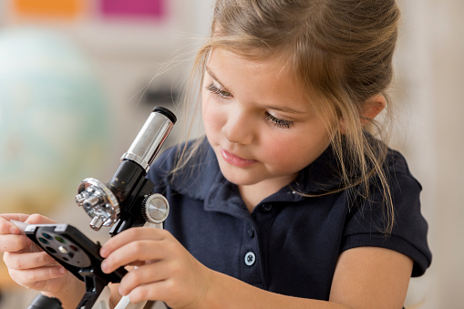 Beautiful Caucasian girl looks at something with a microscope. She attends a STEM elementary school. She is wearing a school uniform and has blond hair.
