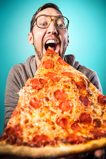 An adult man prepares to take a bite from a humorously large slice of pepperoni pizza, a look of joy and excitement on his face.  Vertical image with cyan / blue background.