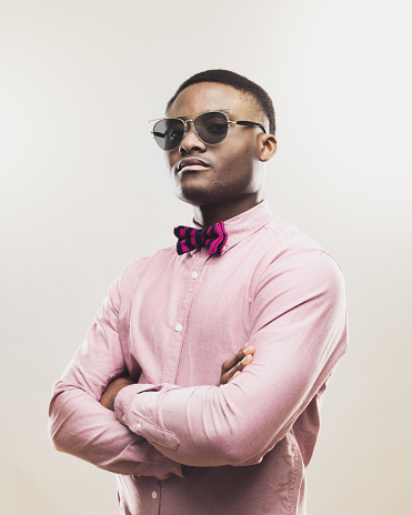 Portrait of young african american man looking at camera with an attitude. Male model wearing bow tie and sunglasses with serious expressions against gray background. Vertical studio photography from a DSLR camera. Sharp focus on eyes.