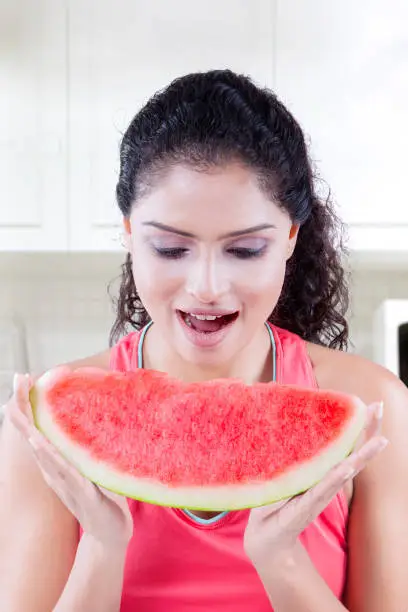 Image of attractive young woman looks happy while eating a big slice of sweet watermelon in the kitchen