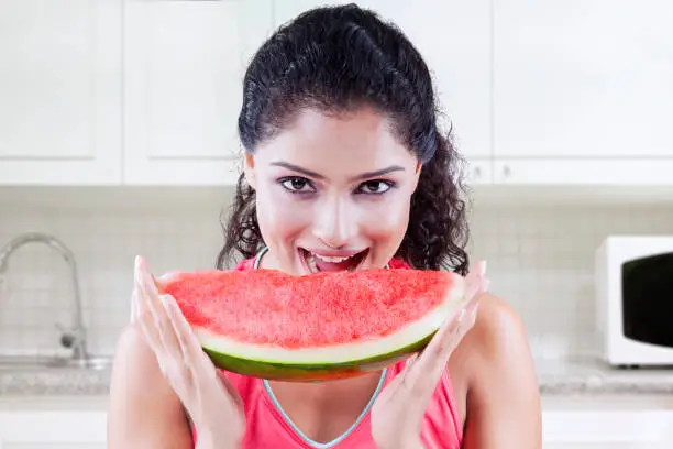Young Indian woman looking at the camera while holding a big slice of sweet watermelon in the kitchen