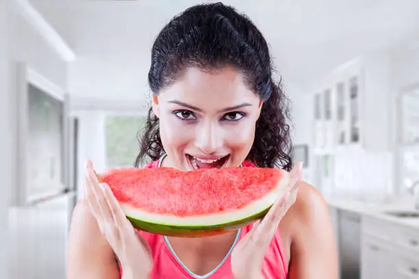 Beautiful young woman looking at the camera while holding a big slice of sweet watermelon at home