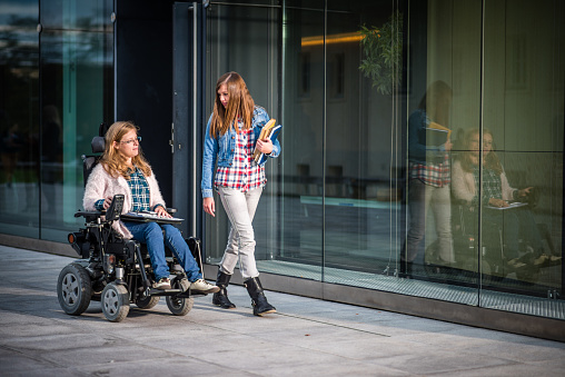 Disabled female student and her friend walking in campus, university building in background.