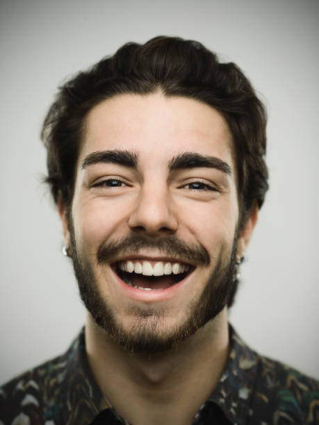 Close-up portrait of a real young confident man. Smiling male is against gray background. He has brown hair and beard. Vertical studio photography from a DSLR camera. Sharp focus on eyes.
