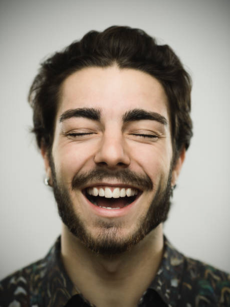 Close-up portrait of a real young confident man. Smiling male is against gray background. He has brown hair and beard. Vertical studio photography from a DSLR camera. Sharp focus on eyes.
