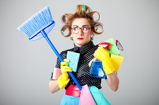 Funny nerdy housewife with curlers in hair wearing yellow protective gloves holding many cleaning products