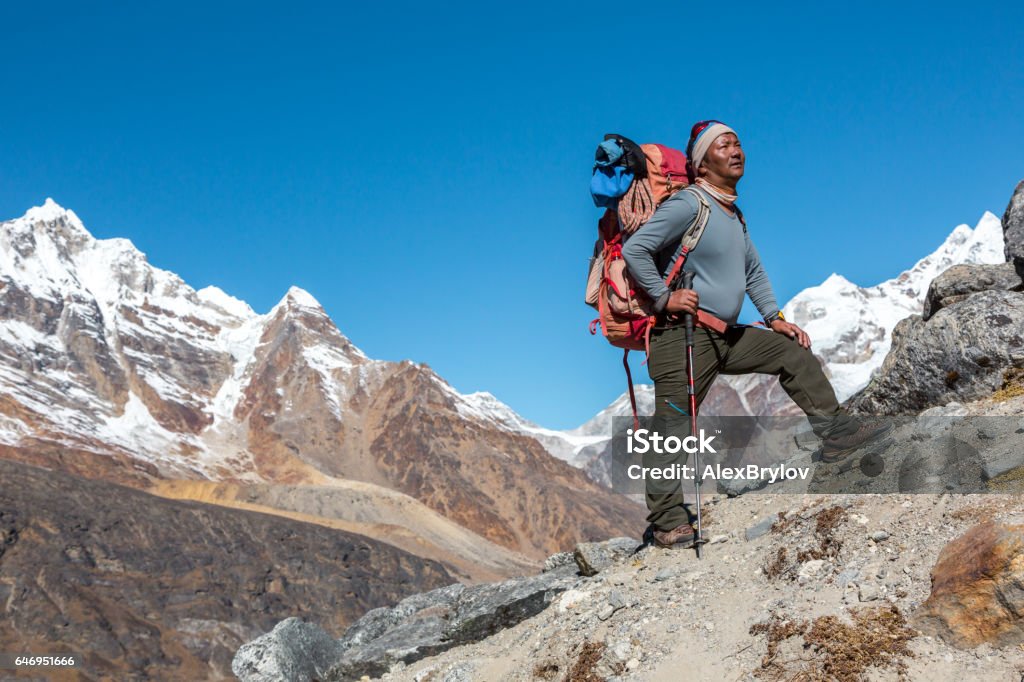 High Altitude Himalaya Nepalese Guide looking Up Mature high Altitude Himalaya Nepalese Mountain Guide staying on rocky Slope with Backpack Climbing Gear and looking Up Sherpa Stock Photo