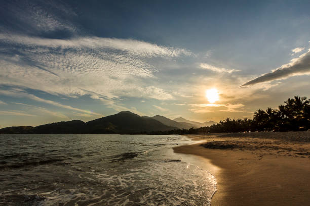 A DSLR Canon photo of a beach in Mangaratiba, Rio de Janeiro, Brazil. The sea is quite calm and there is no one around. The is low in the sky about to set behind the mountains in background.