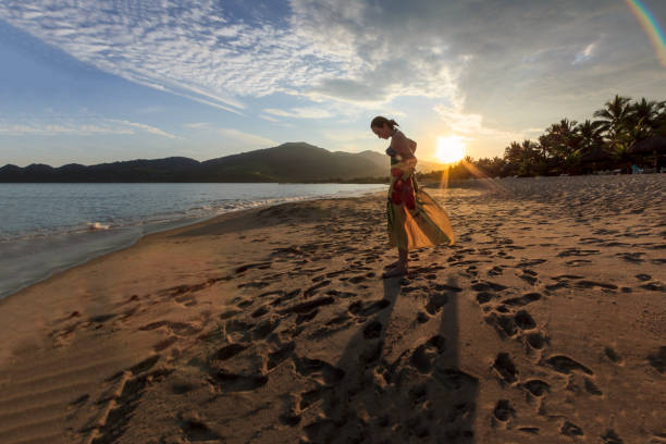 A DSLR Canon photo of a pretty Caucasian Brazilian woman enjoying her vacations on a tranquil deserted beach in Mangaratiba, Rio de Janeiro, Brazil. She is wrapping herself on a beach tower. The sun is just about to hide behind the silhouetted mountains in background. There is no one else around.