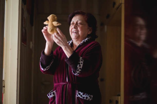 There is one person aged 73. She hold a toy, lookinng at toy and smile. She is nostalgic and childish. She wear a bathrobe, red color.