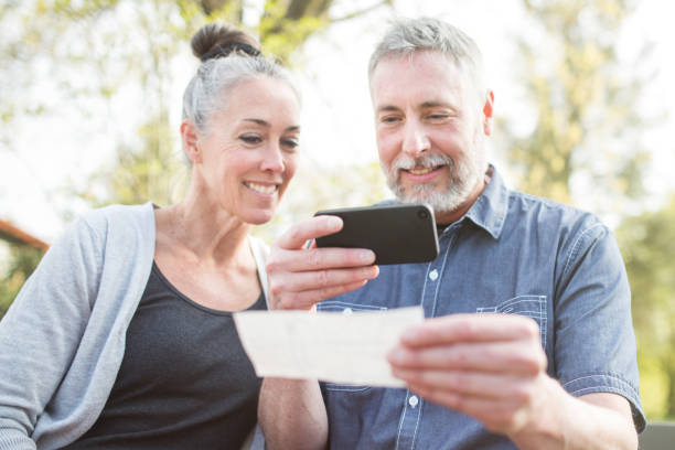 Mature Couple Remote Banking with Phone A gray haired and youthful looking couple in their 50's enjoy time with each other in a beautiful outdoor setting, the sun casting a golden glow on the scene. The man takes a picture of a check with his smart phone for a Remote Deposit Capture to his bank. bank deposit slip stock pictures, royalty-free photos & images