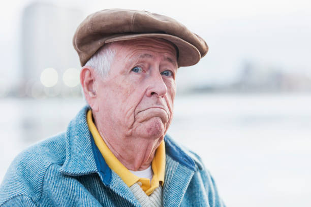 The face of a senior man in his 70s standing on a city waterfront, looking at the camera, frowning.