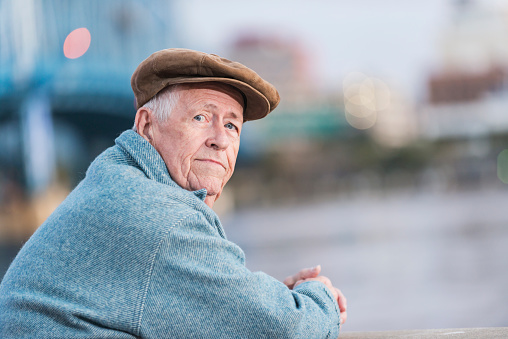 A serious senior man in his 70s wearing a flat cap and coat, on a city waterfront, leaning on the railing. He is looking over his shoulder.