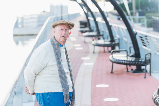 A senior man in his 70s wearing a flat cap, sweater, jeans and a scarf, taking a walk on a pedestrian walkway. He is looking at the camera, hands behind his back, frowning and looking annoyed.
