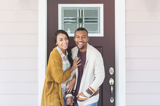 A young couple laughing, standing together in front of the front door of their home. They are wearing preppy, smart casual clothing. The woman is mixed race black and Caucasian and the man is African-American.