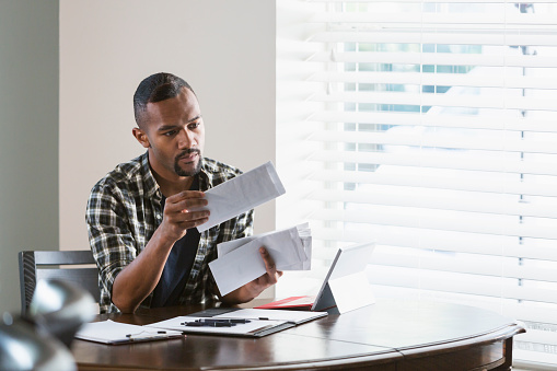 A young African-American man sitting at a desk by a window at home, paying bills. He is looking through a stack of envelopes and has his laptop computer in front of him.