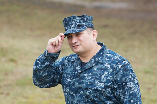An Hispanic man in the US military, wearing camouflage clothing. He is looking at the camera and tipping his hat.