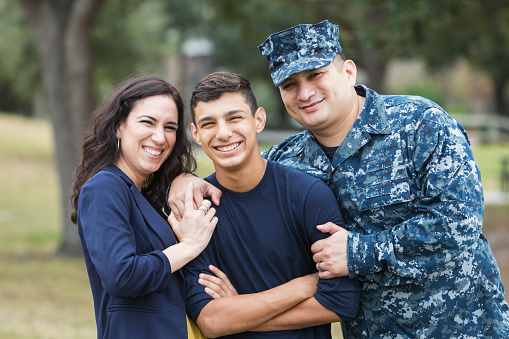 An Hispanic man in the US military, wearing camouflage clothing, standing with his 13 year old son and wife. They are smiling at the camera.