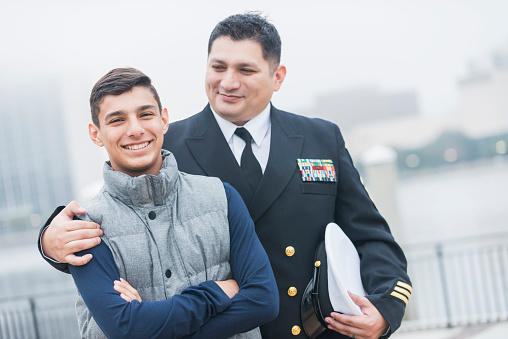 An Hispanic man wearing a military uniform, standing with his teenage son. The 13 year old boy is looking smiling at the camera.