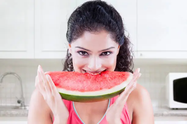 Young Indian woman smiling and looking at the camera while biting a big slice of sweet watermelon in the kitchen