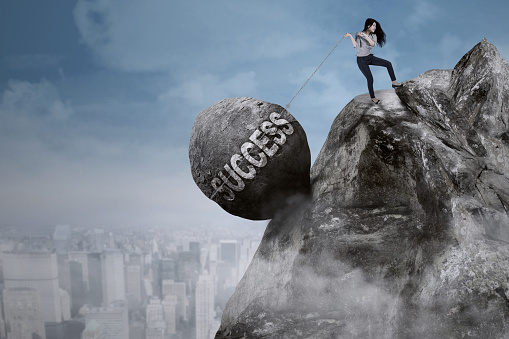 Young Asian businesswoman is pulling big rock with success word while climbing on the cliff