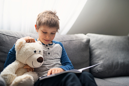 Cute little boy aged 7 reading a book on a couch. The boy is reading to his teddy bear.
