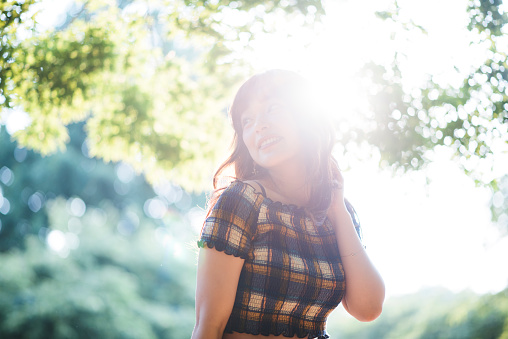 Portrait of a young Japanese woman in Tokyo, Japan. She is smiling, posing and is looking away. Green trees and sun in the back.