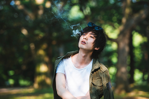 Portrait of a young Japanese man in Tokyo, Japan. He is serious, smoking cigaret, looking at camera and is thoughtful. Green trees in the back.