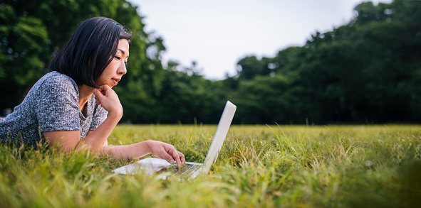 Portrait of young Japanese woman lying on belly on the grass relaxed in park. She is working on laptop. Green trees in the back.