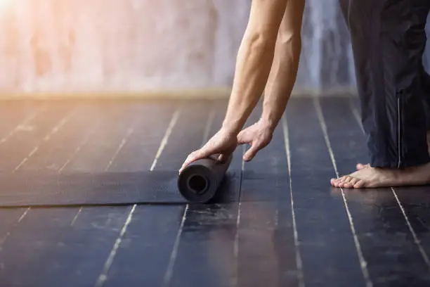 Young yogi men rolling mat after a yoga on black wooden floor