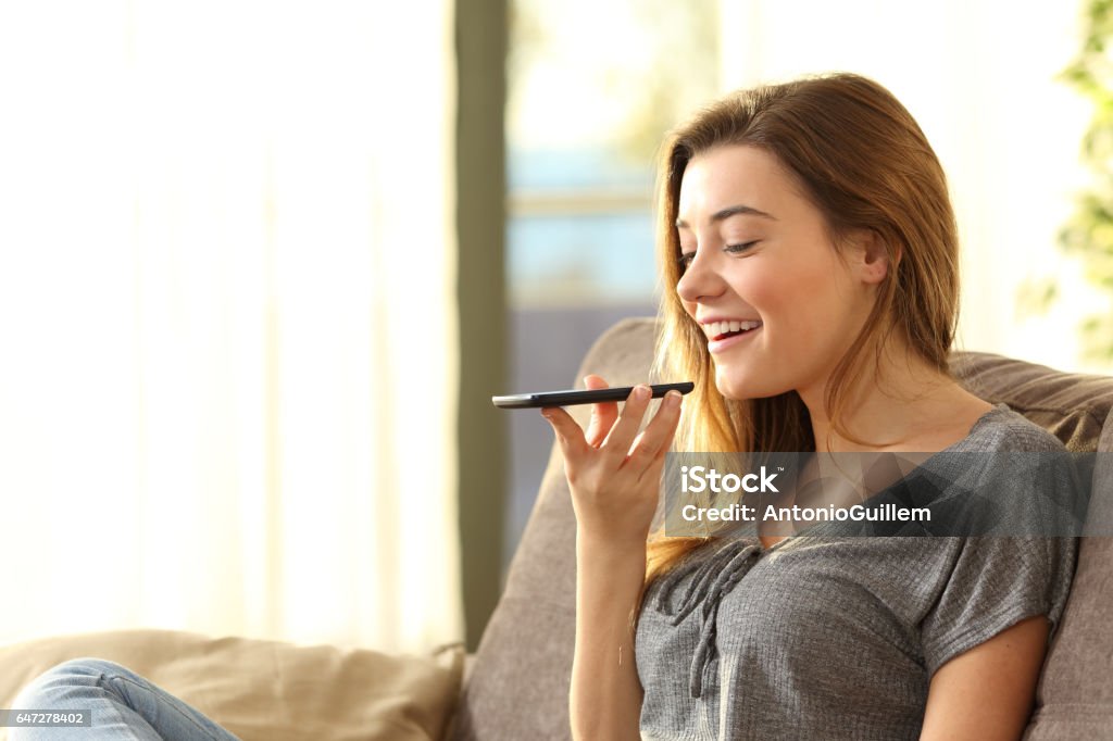 Girl using a smart phone voice recognition Girl using a smart phone voice recognition on line sitting on a sofa in the living room at home with a warm light and a window in the background Mobile Phone Stock Photo