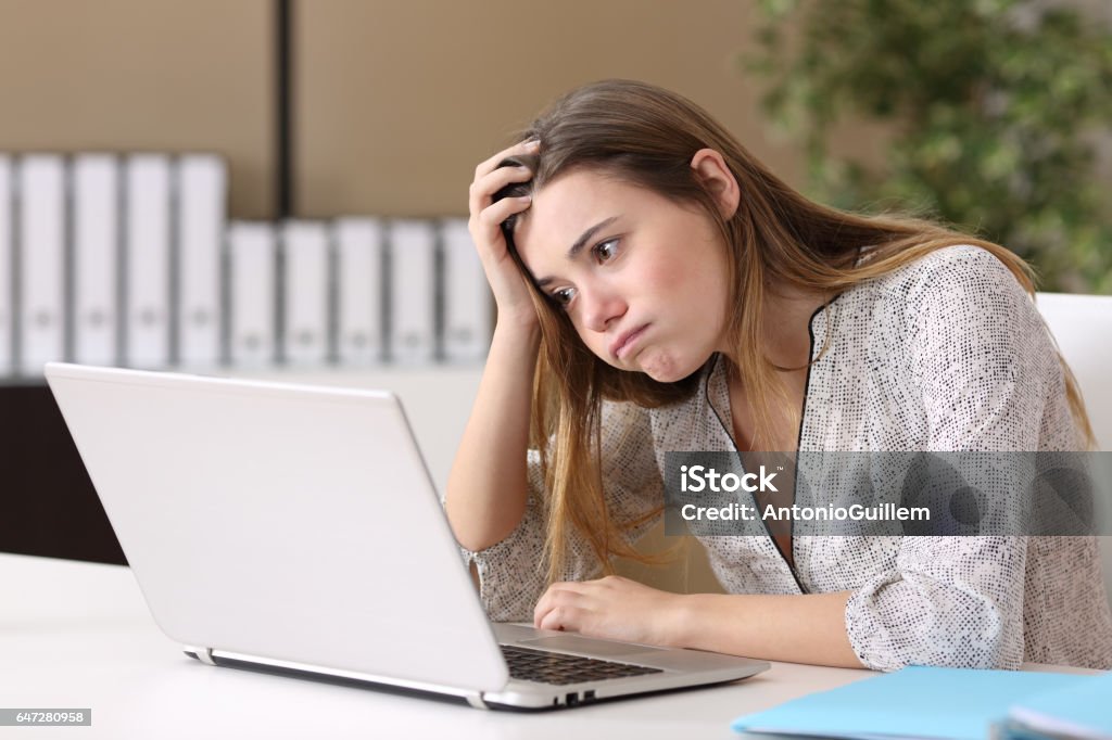 Frustrated intern working on line at office Frustrated intern trying to work on line with a laptop in a difficult assignment in a desktop with an office background Occupation Stock Photo