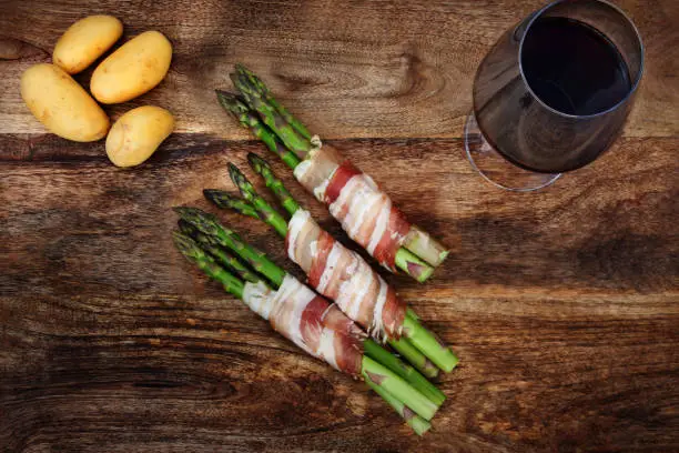 Green asparagus in bacon with potatoes and a glass of wine on a rustic wooden table