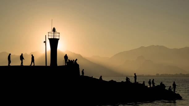 Silhouette of lighthouse in Antalya marina at sunset.The silhouettes of walking, fishing, sitting people are seen.The sun is hidden right behind lighthouse.