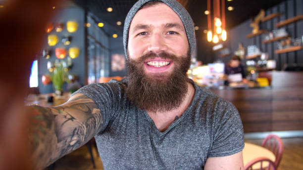 Bearded and tattooed hipster taking a selfie in the cafe.