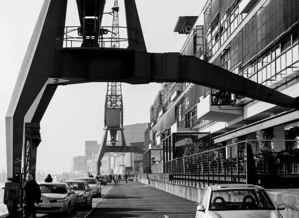Hamburg, Germany - Februar 15, 2017: Historic harbor cranes next to modern architecture on the river promenade in Altona. People are walking by. The picture is monochrome.