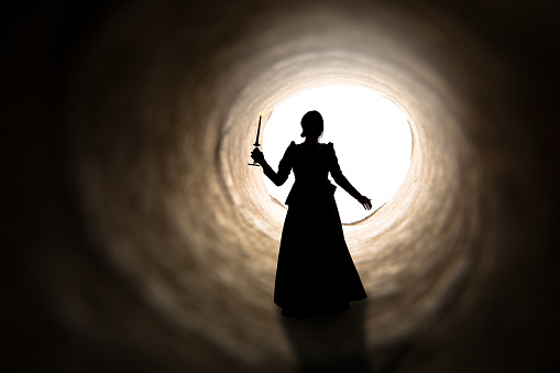 A silhouette of a woman in vintage dress holding a candle as she walks toward the light at the end of a tunnel.