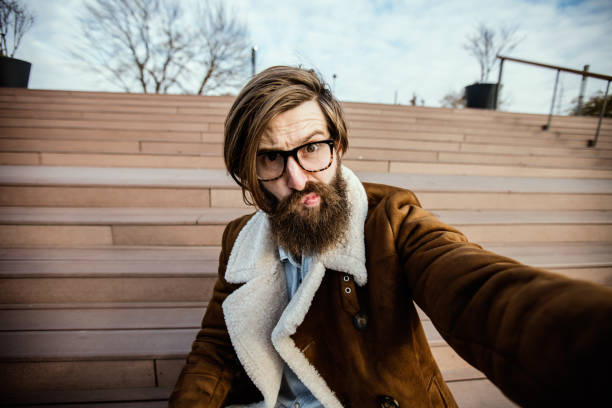 chico pelo largo sentado en una escalera haciendo cara tonto y tomar foto selfie - stubble men tattoo sensuality fotografías e imágenes de stock