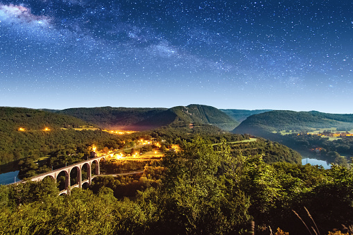 High angle view landscape of Bugey mountains, from beginning of Jura in France, Auvergne-Rhone-Alpes region. There is an old stone viaduct from Cize-Bolozon villages crossing Ain river in the valley. Arch bridge architecture under bright full moon, moonlight in summer with beautiful blue clear star sky, with milky way from space. This viaduct is a combination rail and vehicular, connecting the communes of Cize and Bolozon. An original span built in the same location in 1875 was destroyed in World War II. Reconstructed as an urgent post-war project due to its position on a main line to Paris, the new viaduct reopened in May 1950. It carries road and rail traffic at different levels.